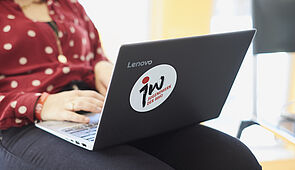 Auf dem Bild ist eine junge Frau zu sehen, die sitzend an einem Laptop arbeitet. Auf diesem ist ein Sticker mit dem Logo des Jugendwerks.